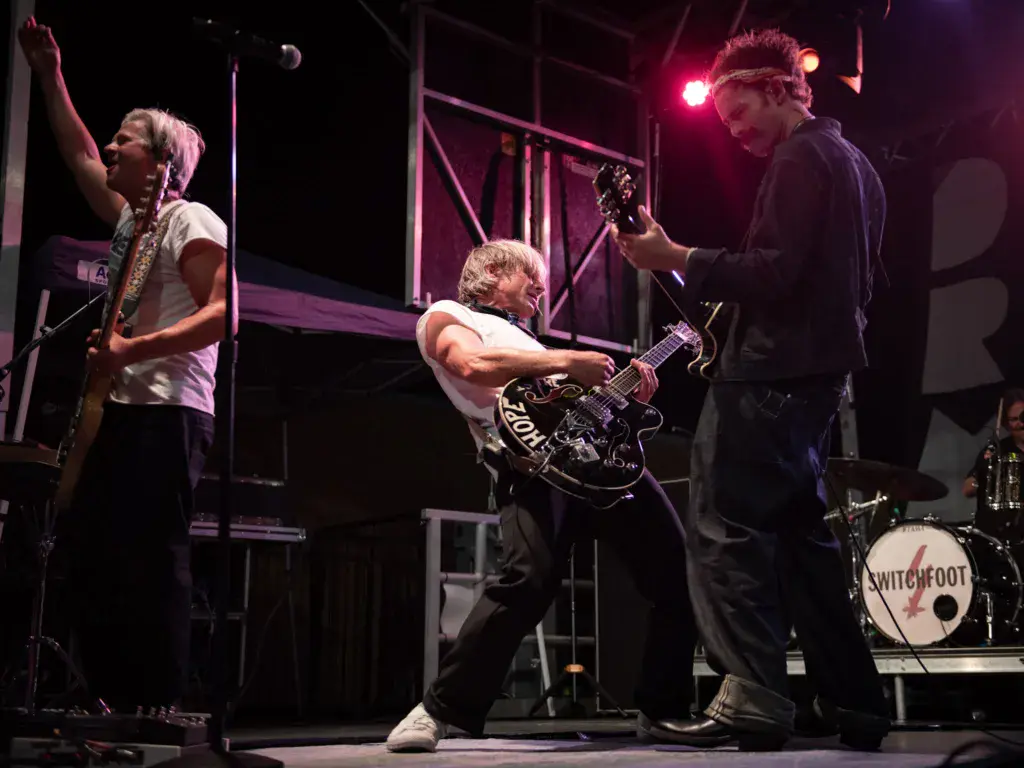 Band performing on stage with three musicians playing guitars and drums under pink stage lighting