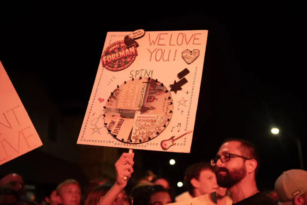 Audience member holds a handmade sign reading 'WE LOVE YOU!' at an outdoor night concert.