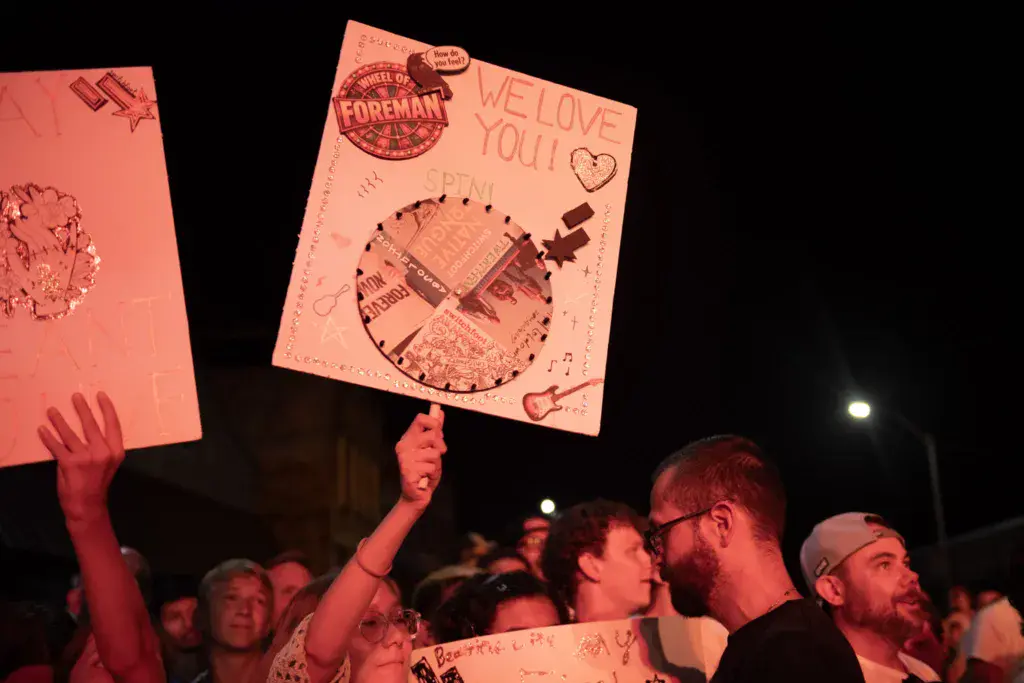 Crowd members holding pink handmade signs with messages at a nighttime festival event.