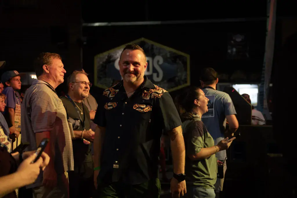 A man in a black embroidered western shirt stands among festival attendees at night near stage signage.