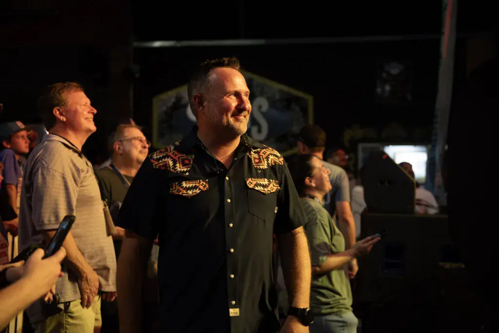 A man in a black shirt with gold embroidery stands among festival attendees in an outdoor evening setting.