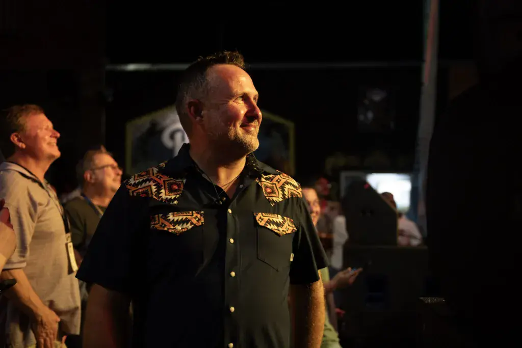 A man in a black embroidered Western shirt stands among festival attendees at night.