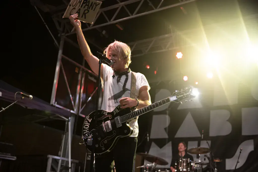 A guitarist performs on stage holding a sign above their head while playing a black electric guitar under bright stage lights.
