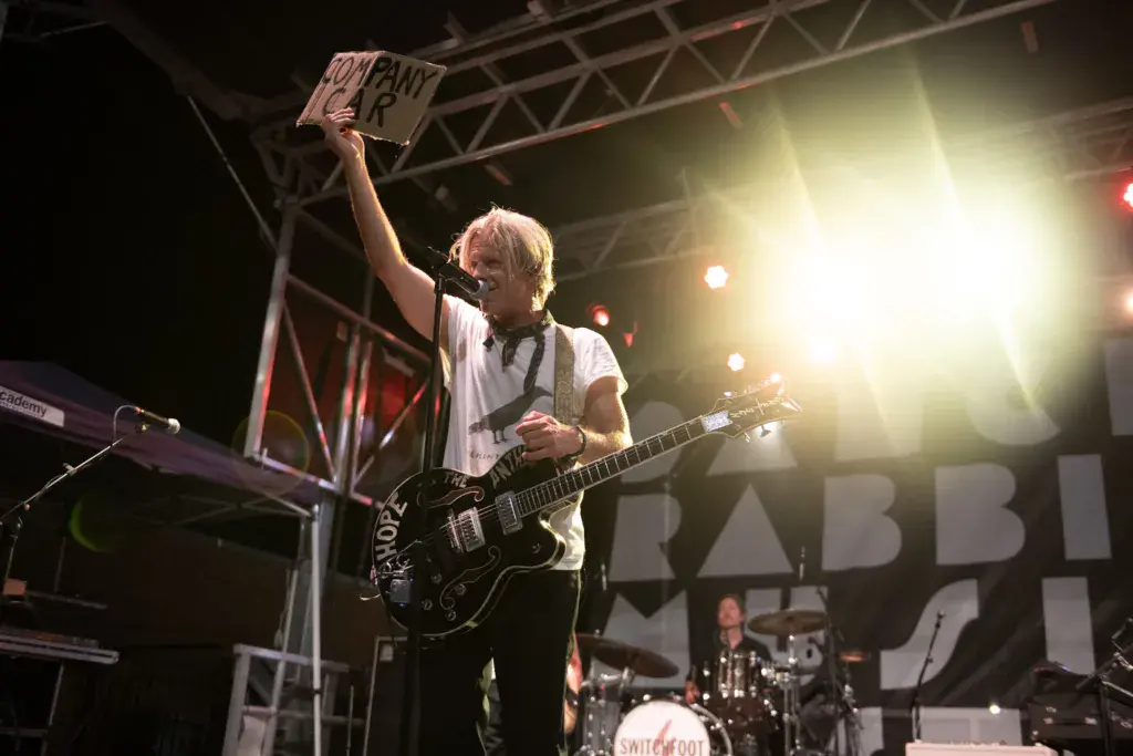 Guitarist performs on stage holding a sign aloft while playing a black electric guitar under bright stage lights.