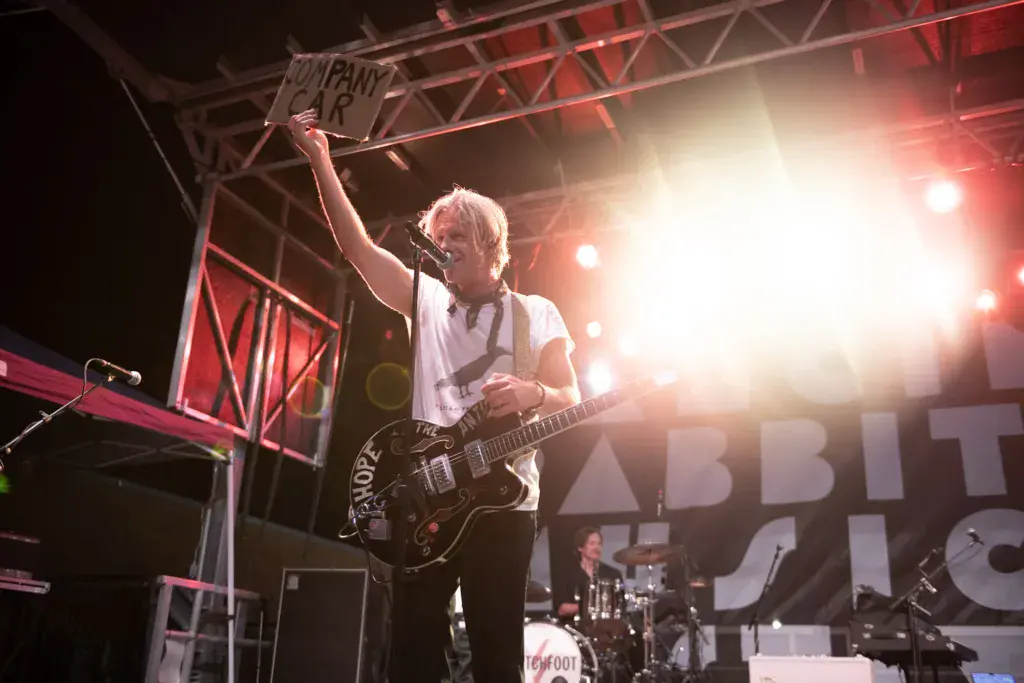 A guitarist holds a sign aloft while performing on stage under bright red and white lights.