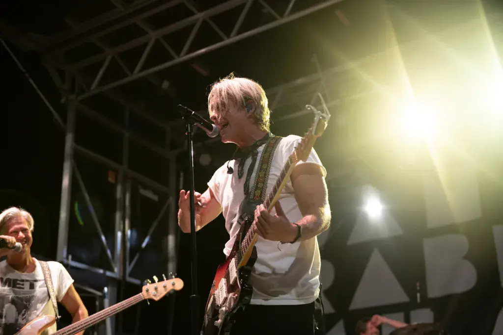 A musician with blonde hair plays an electric guitar and sings into a microphone on stage during an evening performance.