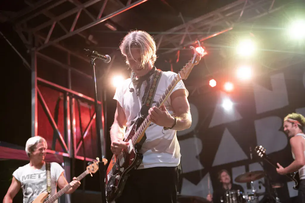 A guitarist with blonde hair performs on stage under dramatic red and white stage lighting at a music festival.