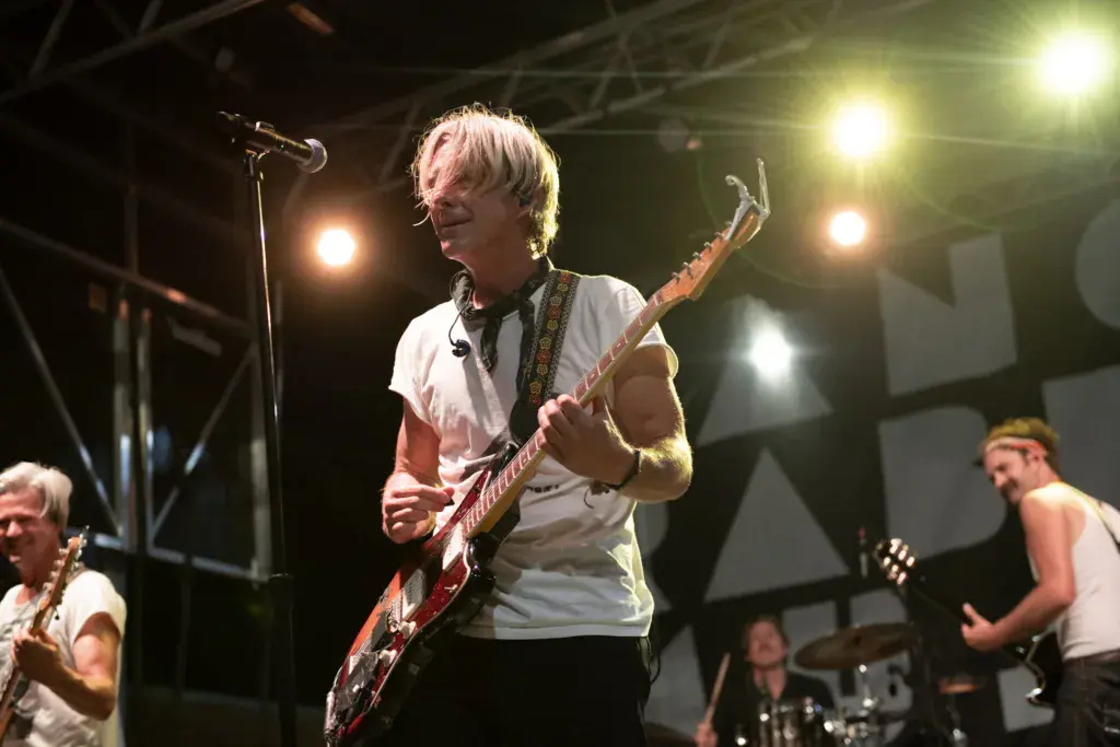 A guitarist with blonde hair performs on stage under bright stage lights while band members play alongside.