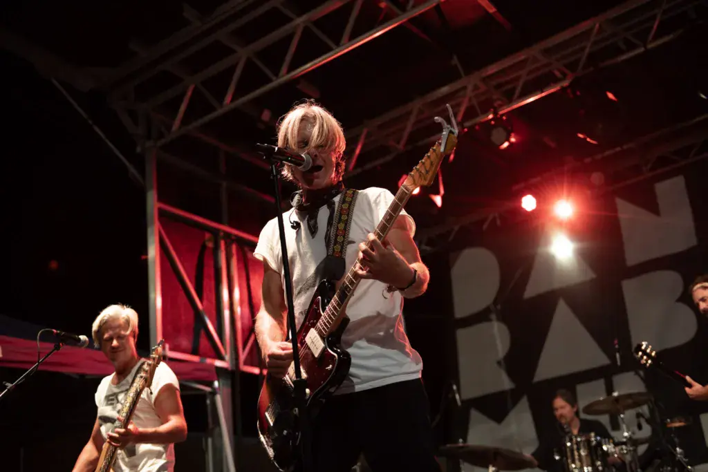 A guitarist in a white t-shirt performs on stage under red stage lighting while a saxophonist plays alongside.