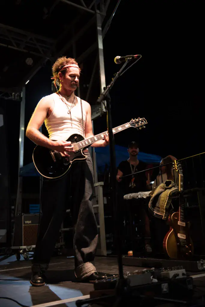 Guitarist with curly hair performs on stage under bright lights, wearing a white tank top.