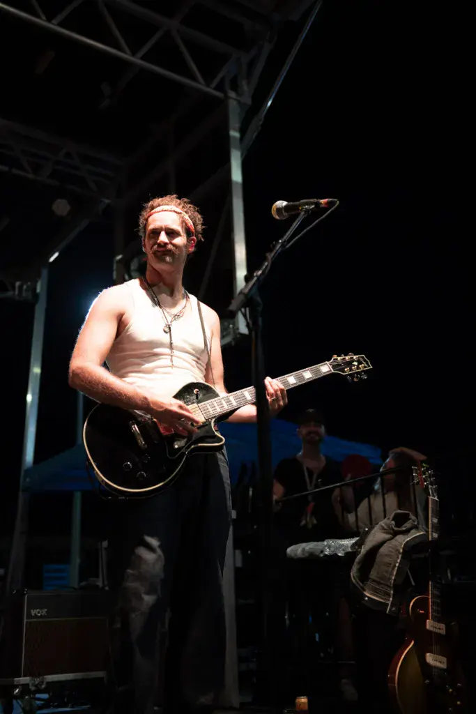 A guitarist performs on stage at an outdoor music festival, wearing glasses and a white sleeveless shirt while playing a black electric guitar.