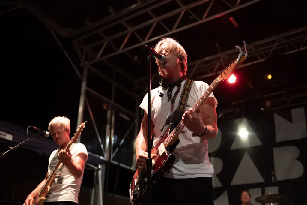 Two musicians perform on stage with guitars under red and white stage lighting at night.