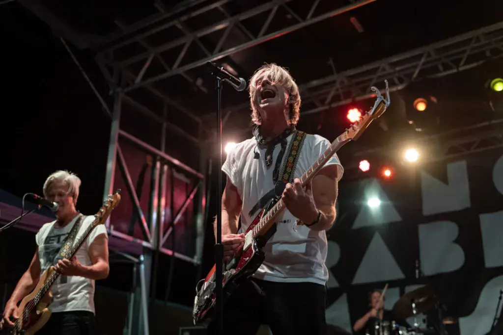 Band performing on stage at night with guitarist singing at microphone and bassist on left under stage lighting.