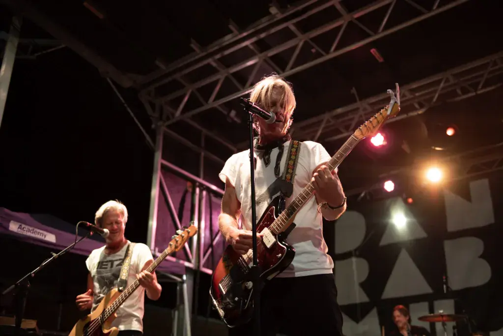 Two musicians perform on stage with guitars under bright lights at an outdoor festival venue.