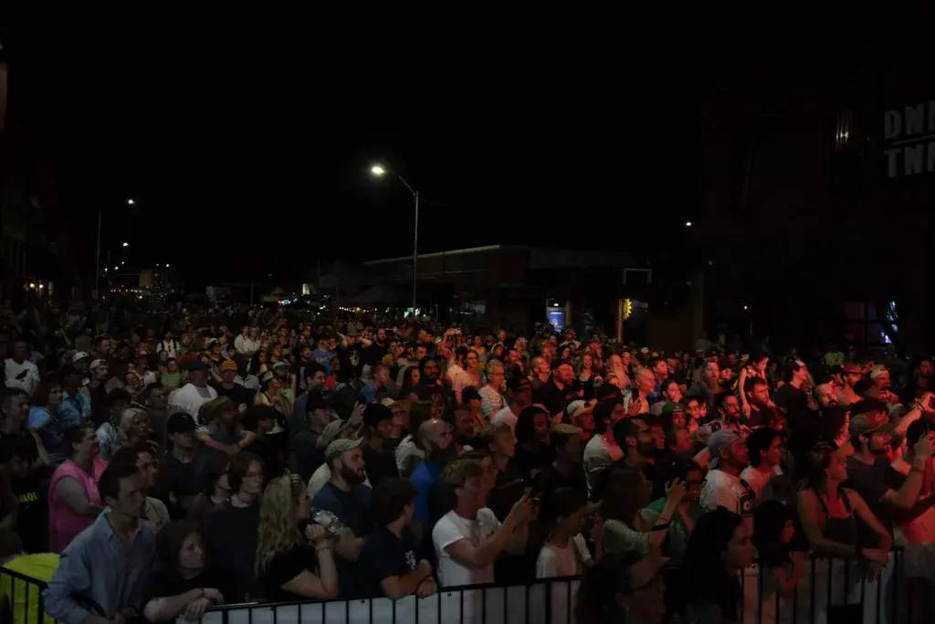 A large crowd of festival-goers gathered at night, illuminated by red stage lighting and overhead street lights.