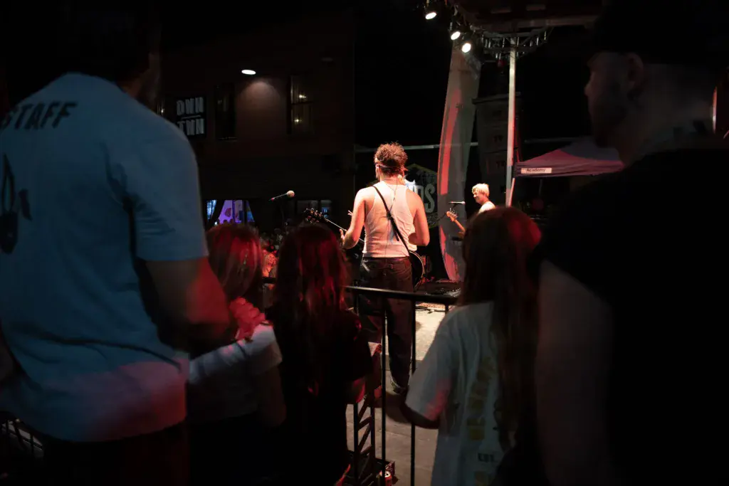 Crowd watches a performer with a guitar on an illuminated stage at an evening music festival.