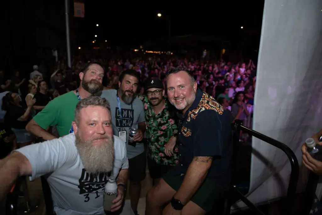 A group of five men pose together at night in front of a large crowd at an outdoor music festival.