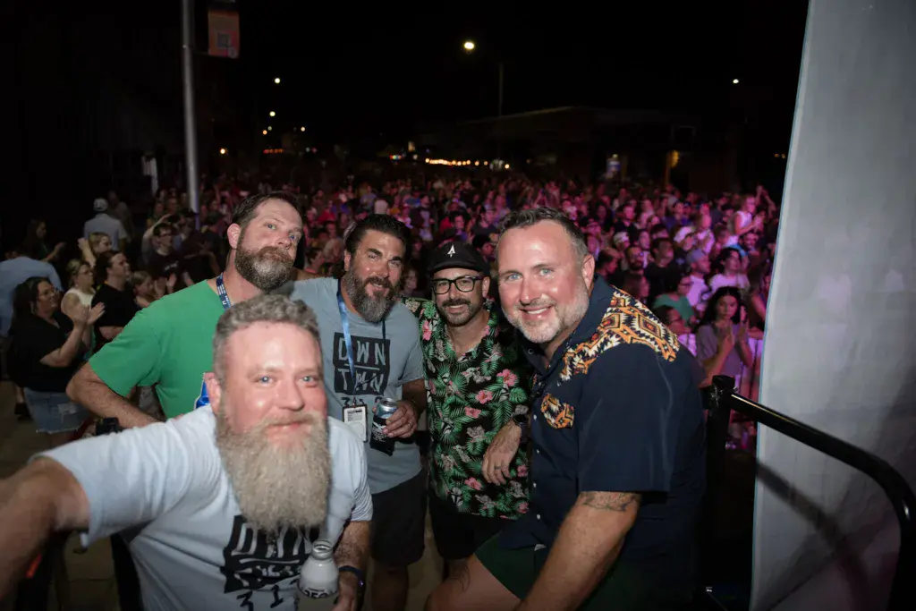 Five men pose together for a photo at a nighttime outdoor festival with a large crowd in the background.