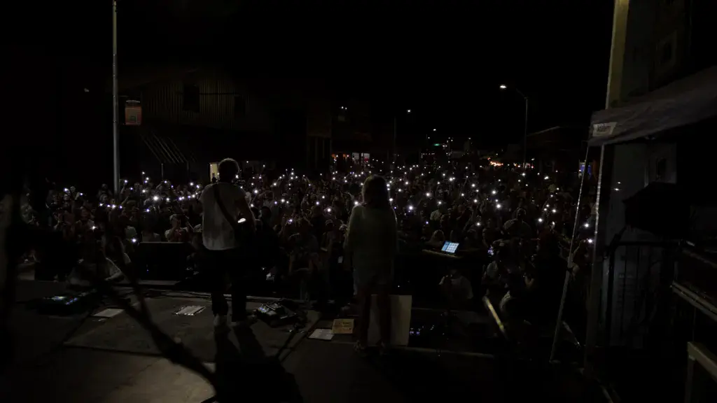 A large crowd at night holds up illuminated phones and lighters during a concert performance at an outdoor festival venue.