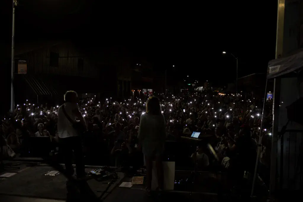 Large nighttime crowd at outdoor festival holding up illuminated phones while performers stand on stage.