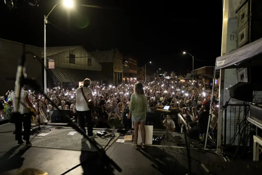A large nighttime crowd fills a downtown street with their phones raised, watching performers on an outdoor stage.