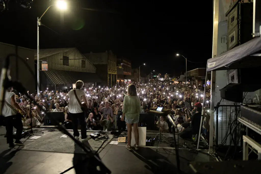 Performers on an outdoor stage with a large crowd holding up phones at night during a music festival on a city street.