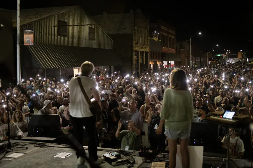 Performers on stage facing a large crowd with cellphones raised during a nighttime outdoor music festival.