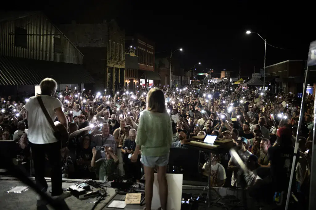 Performers on stage at night with a large crowd holding up illuminated phones during an outdoor music festival.