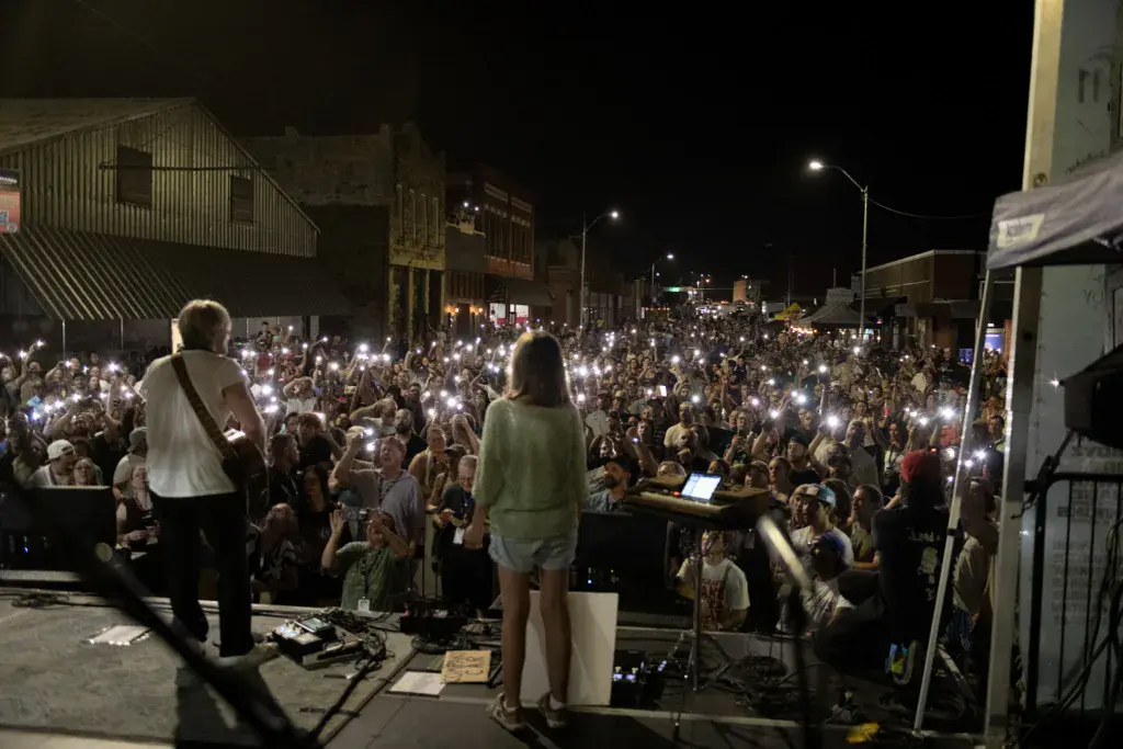 Two performers on stage at night facing a large crowd holding up lit smartphones during a festival performance.