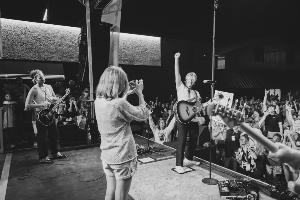 Band performing on stage at night with enthusiastic crowd raising hands and phones to record.