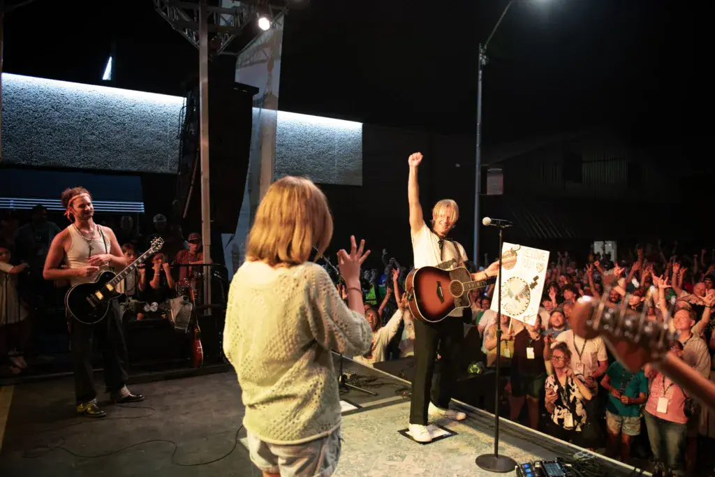 Performers on an outdoor stage with a large crowd of festival attendees dancing and raising their hands in front of them at night.