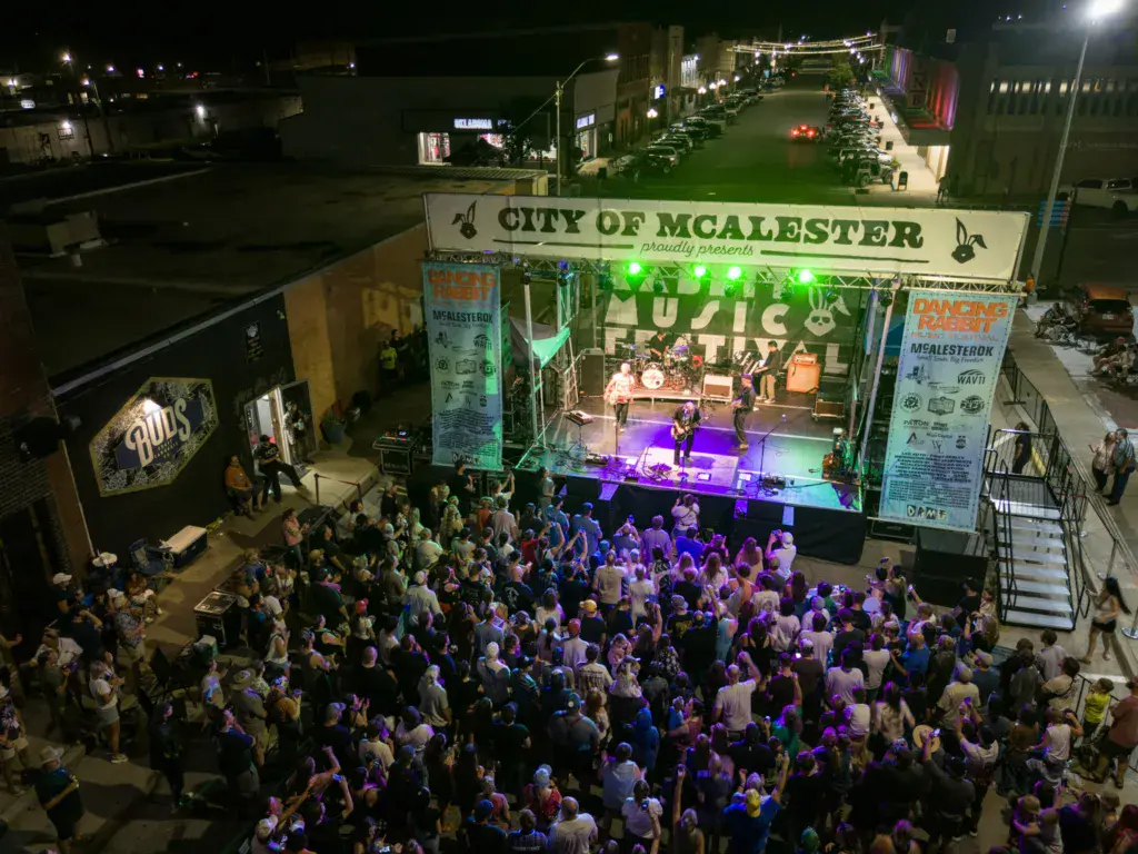 Overhead view of a large crowd gathered in front of an illuminated stage at night with City of McAlester signage.