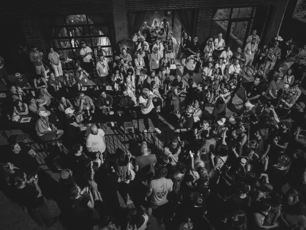 Black and white overhead view of a large crowd gathered inside an industrial brick venue around a central performance space.