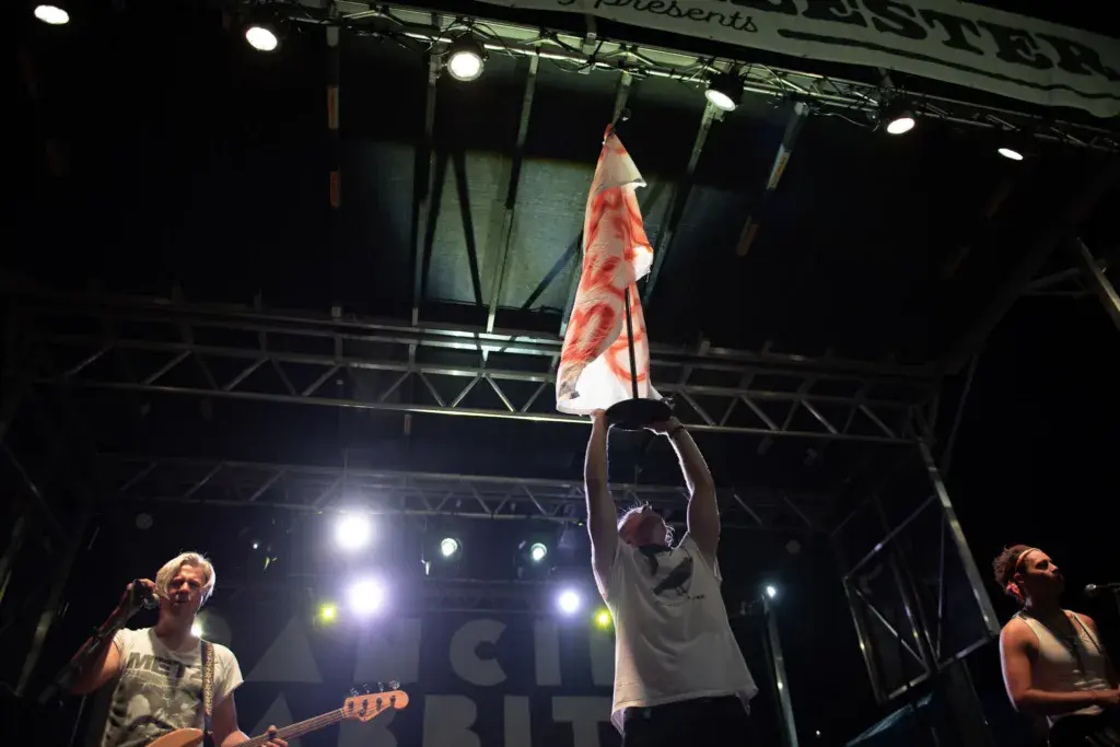 A performer on stage waves a red and white flag while band members play instruments under bright stage lights.