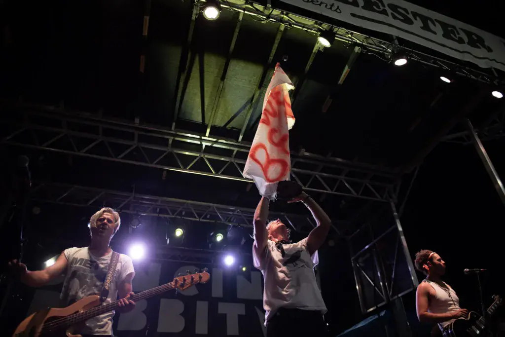 A band performs on stage at night with one member holding up a red and white flag during the set.