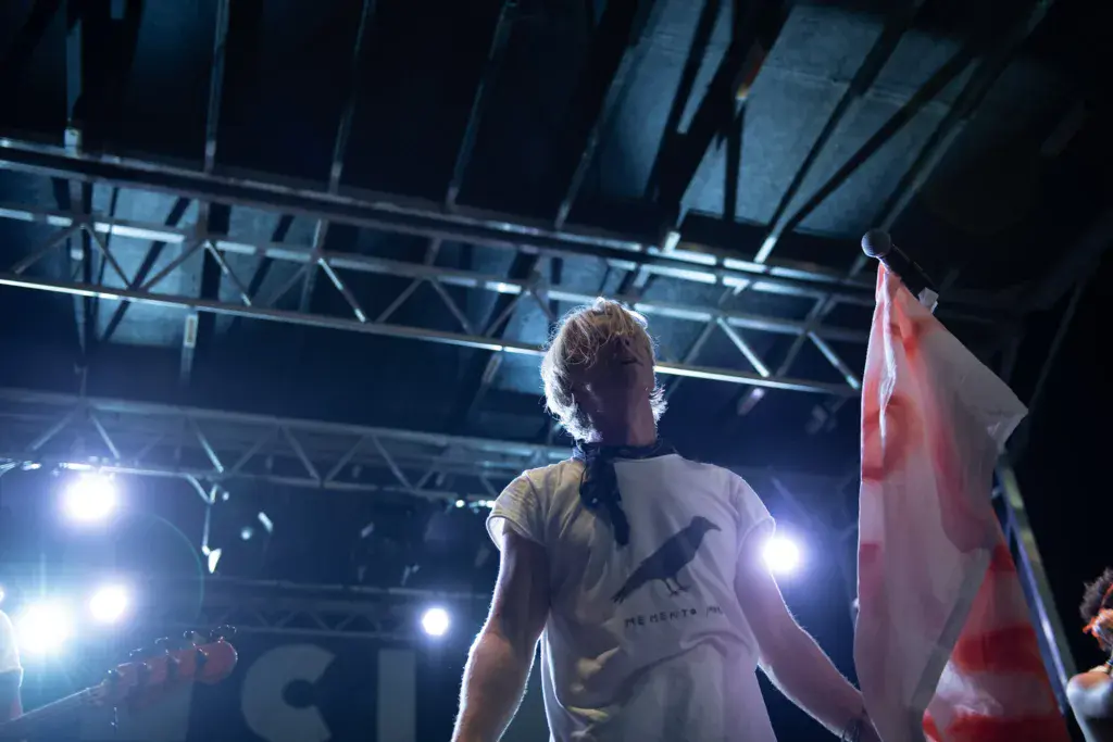 A performer on stage under bright lights holds a flag while addressing the crowd at night.