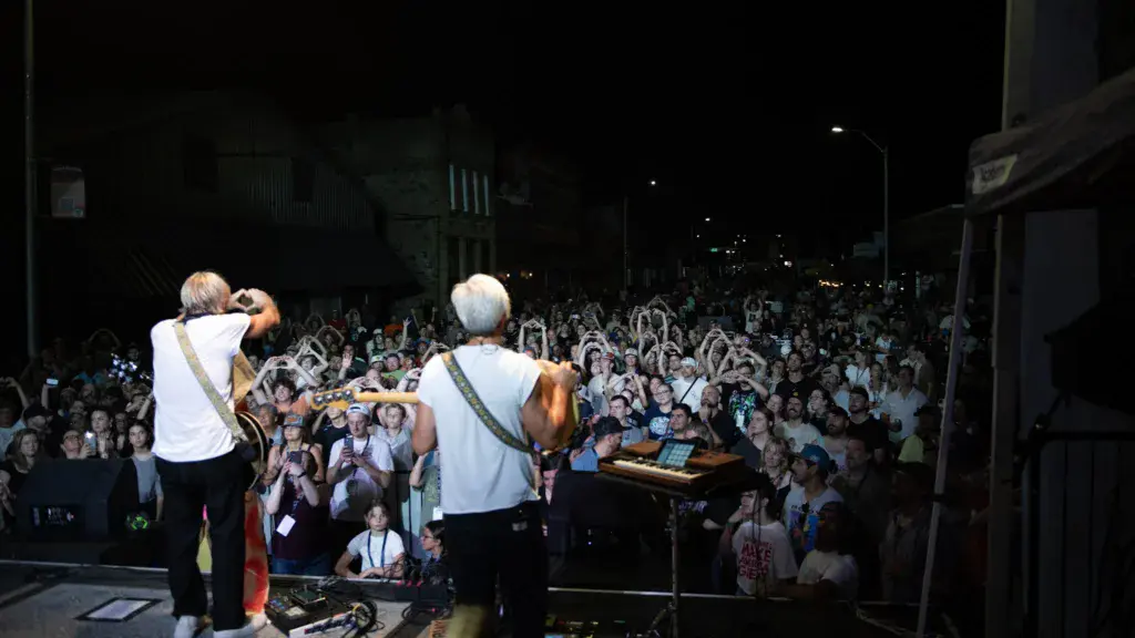 Two performers on stage with guitars face a large crowd under nighttime festival lighting.