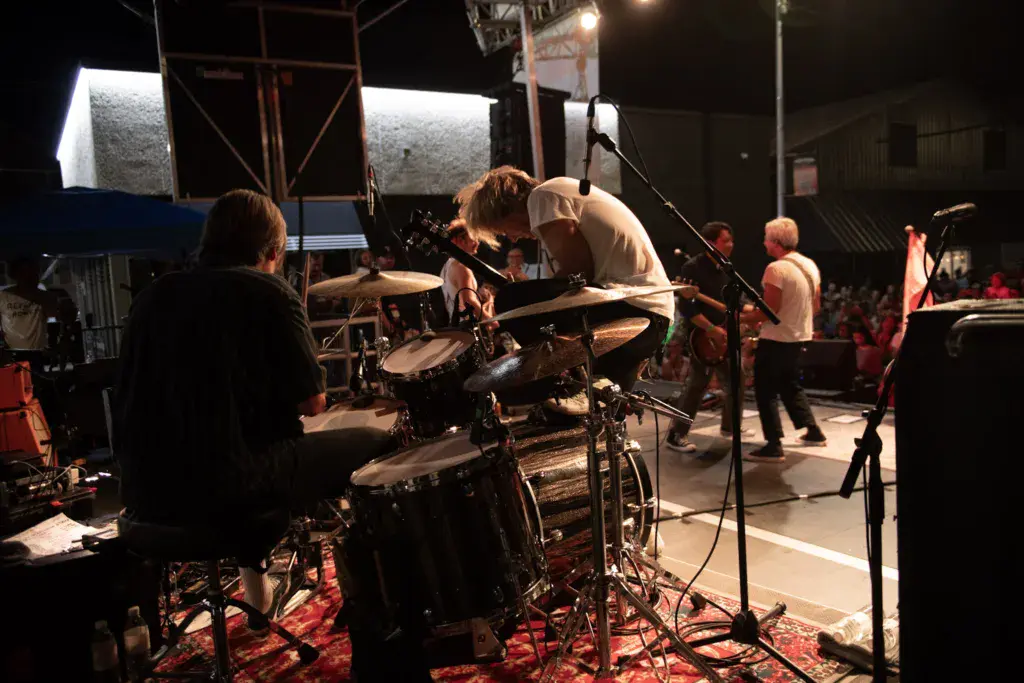 Band performing on outdoor stage at night with drummer in center and crowd watching from below.