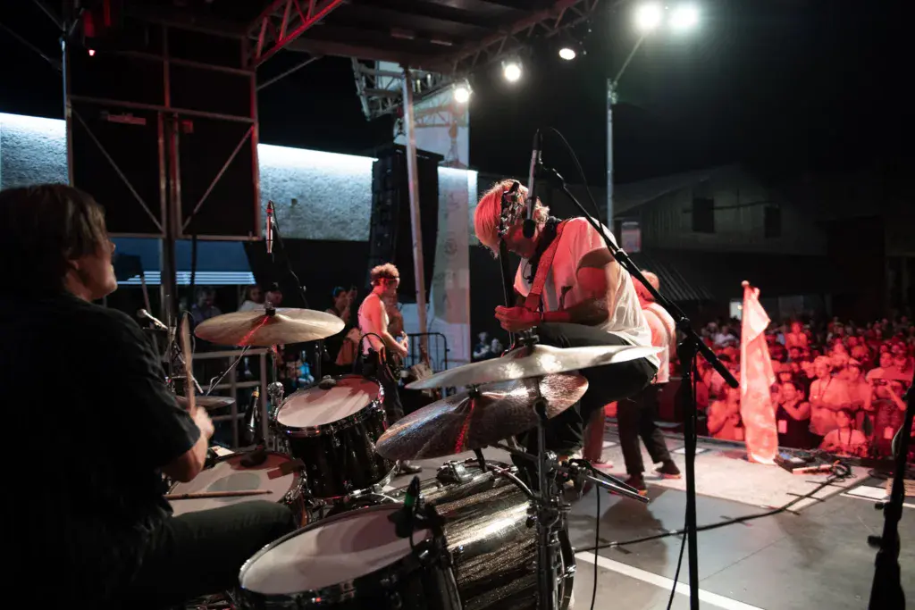 A drummer performs on stage at an outdoor music festival with a large crowd watching under stage lights at night.