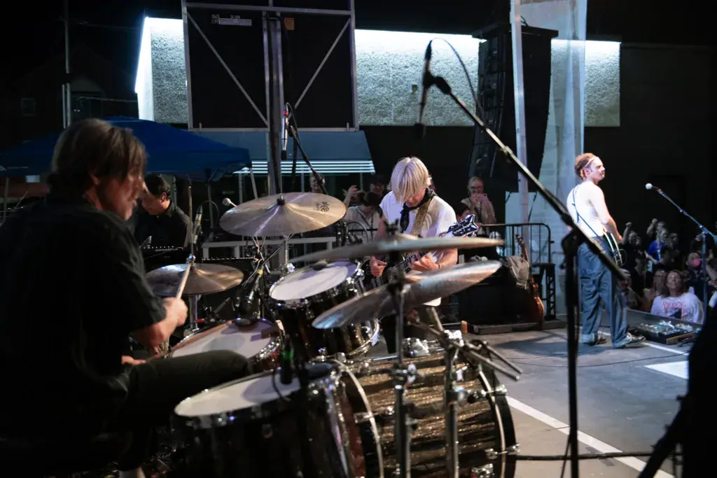 A band performs on an outdoor stage at night, with a drummer in the foreground and spectators visible in the crowd.