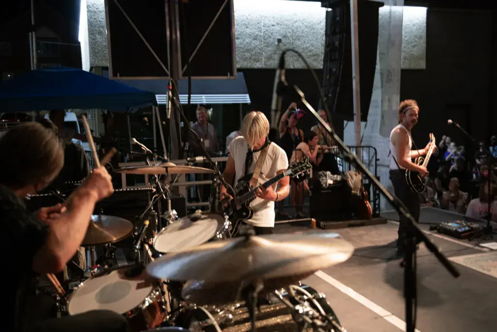 A band performs on an outdoor stage with drummers in the foreground and guitarists visible, audience members watching from the side.
