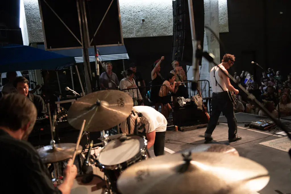 A band performs on an outdoor festival stage at night with a drummer in the foreground and crowd visible in the background.