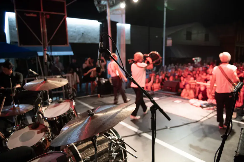 A live band performs on stage at night with a large crowd gathered in front, illuminated by red stage lighting.