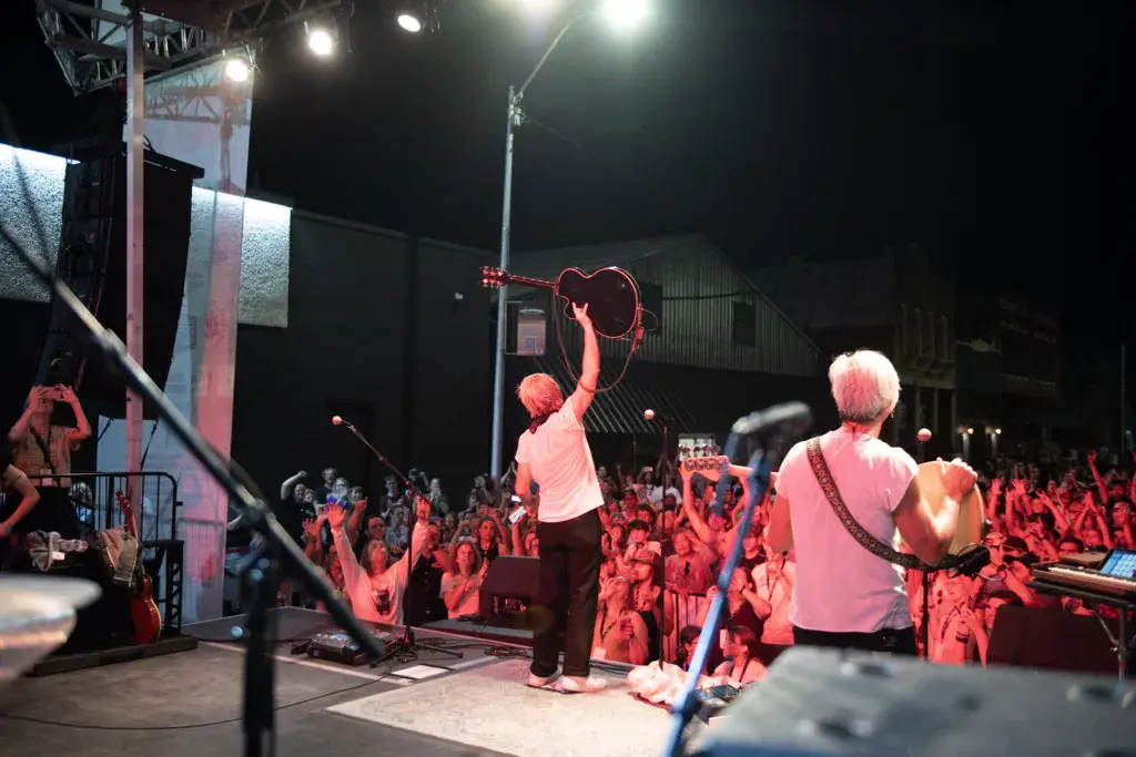 A performer with a guitar raises their arm on stage as a large crowd watches under stage lights at night.