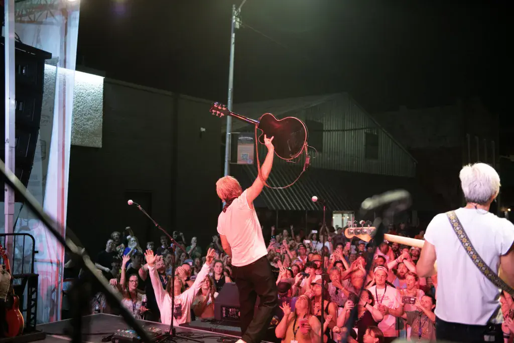 A performer holds up an acoustic guitar on stage while a large crowd watches under red stage lighting at night.