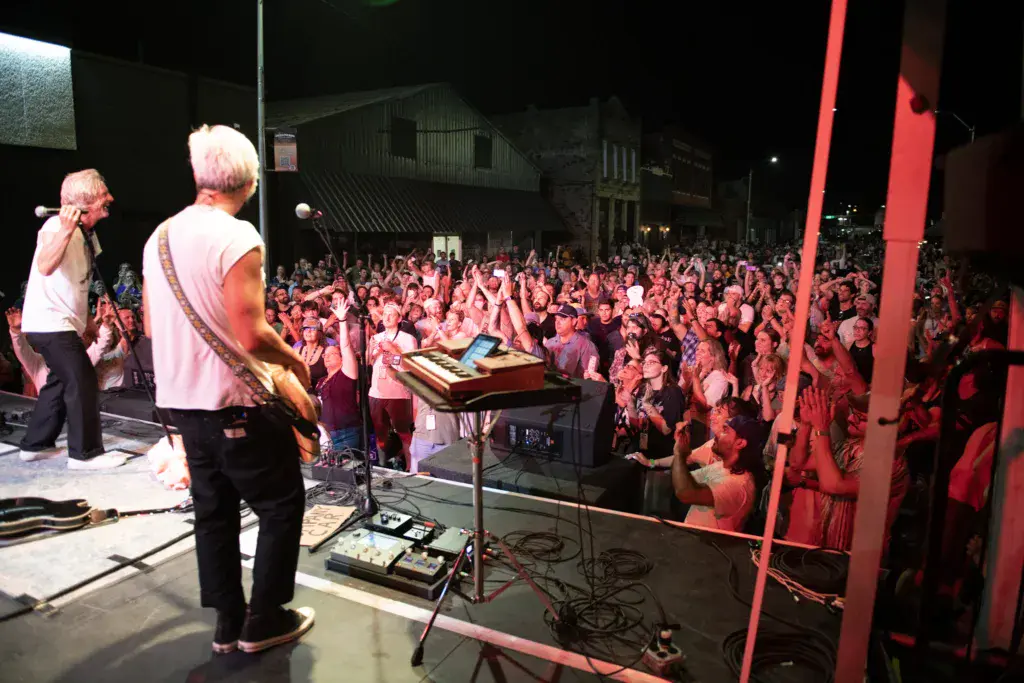 Musicians perform on stage with keyboard and guitar as a large crowd watches under red stage lighting at night.