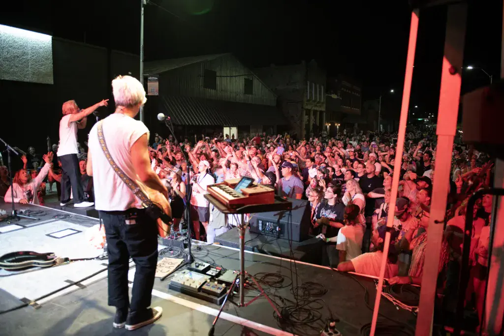 Musicians perform on a stage bathed in red lighting before a large crowd at an outdoor music festival venue at night.