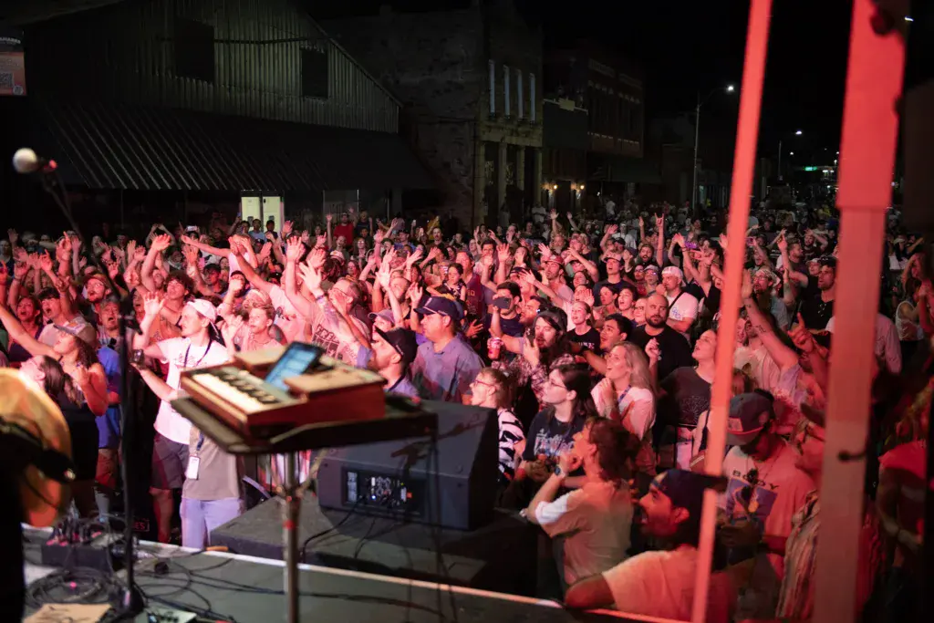 A large crowd with hands raised dances under pink lighting at an outdoor nighttime music festival venue.