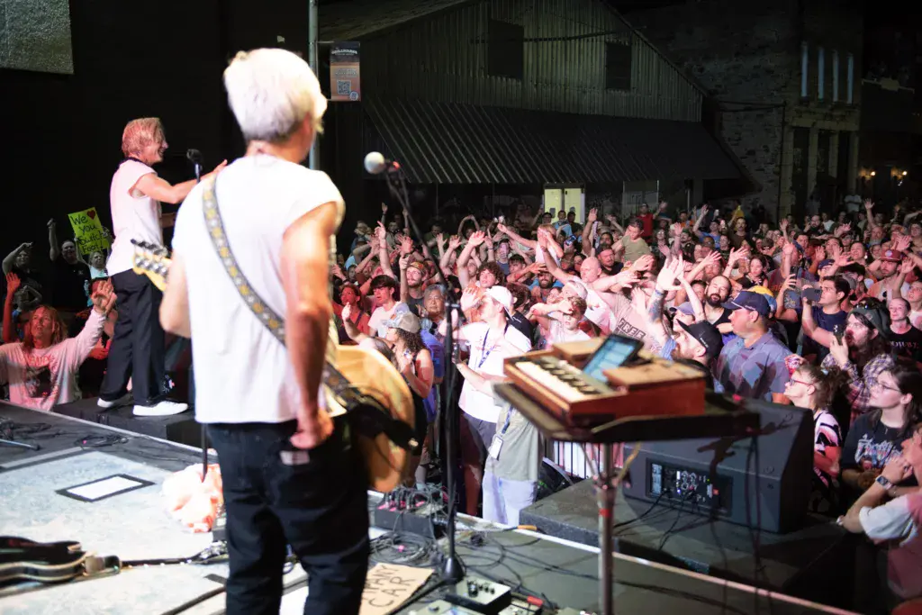 Musicians perform on stage before a large enthusiastic crowd at an outdoor music festival venue.