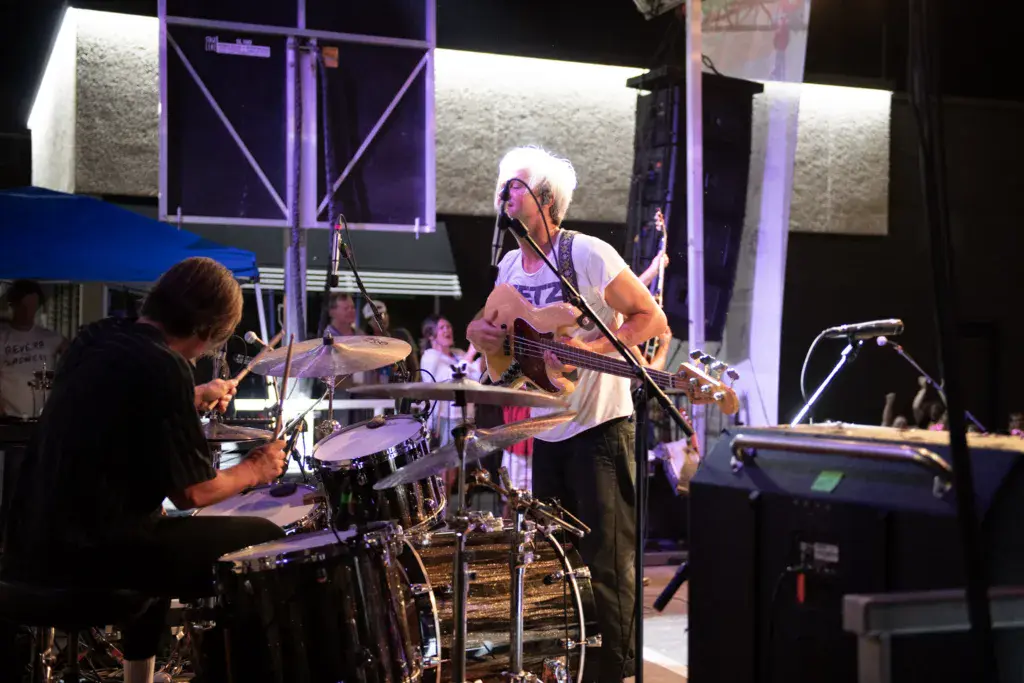 A band performs on an outdoor stage with purple lighting, drummer on left and guitarist-vocalist on right.